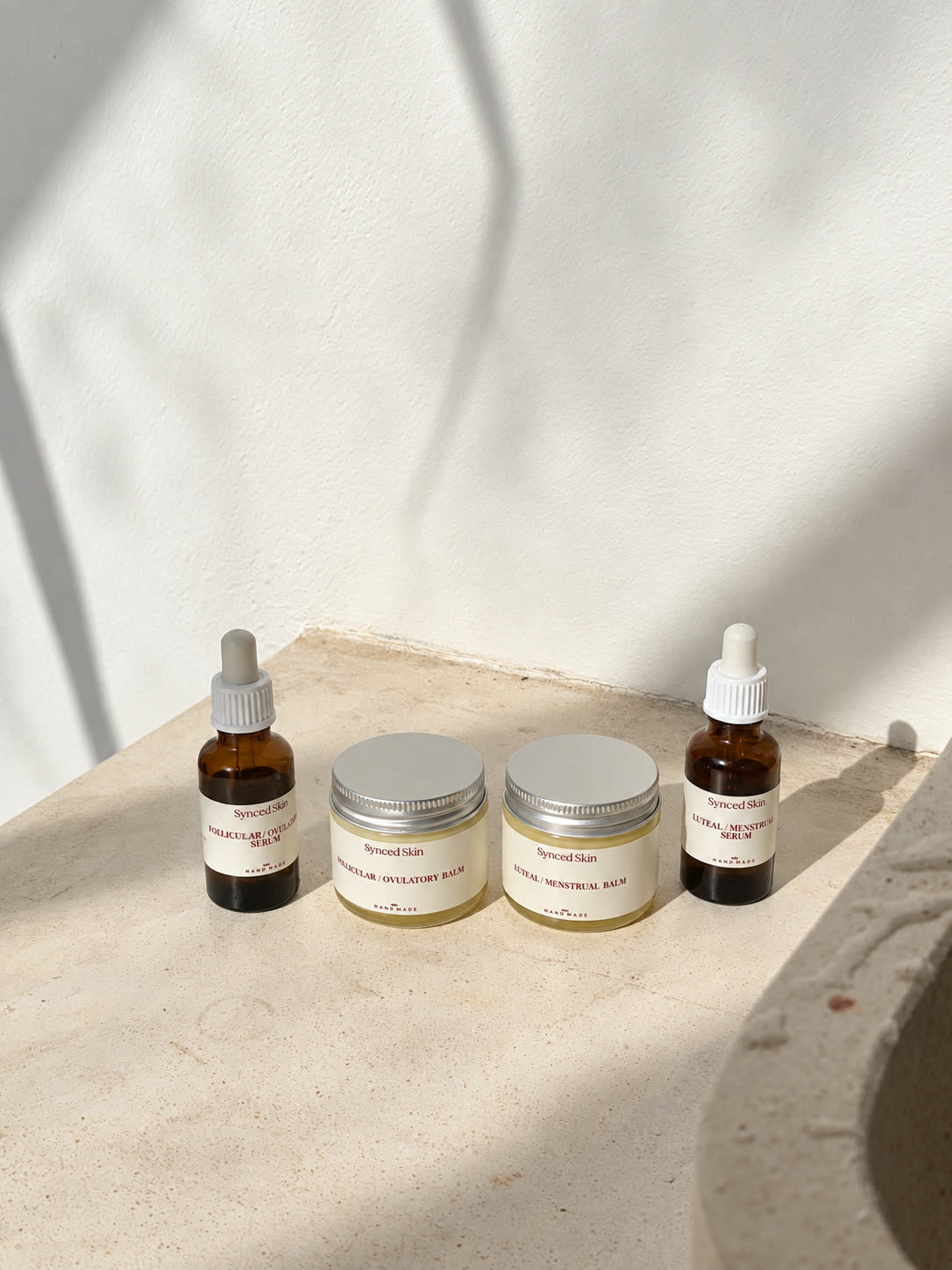 Two Synced Skin face serums and two tallow balms in sunlight on a stone ledge