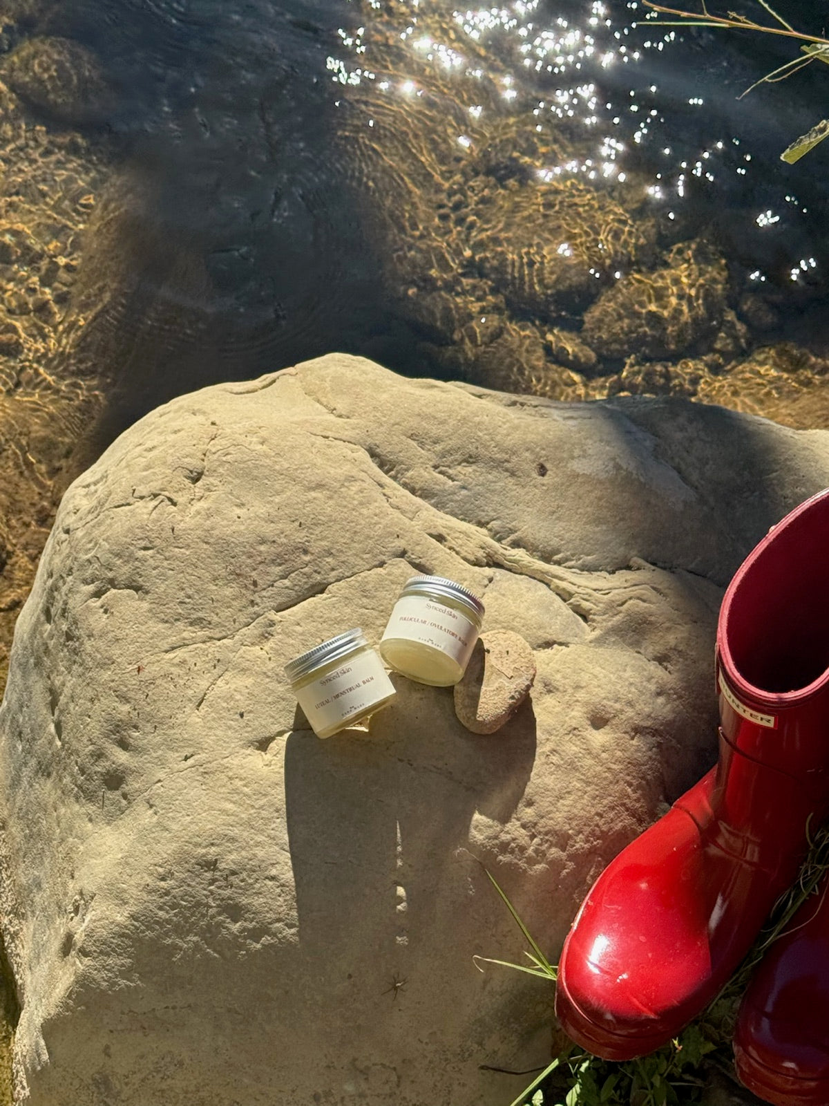Two Synced Skin balm jars on a rock beside a creek and red boots.