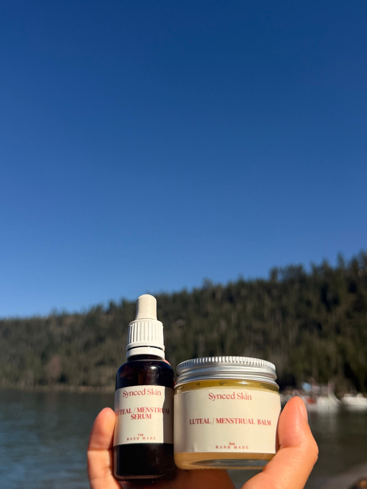 A Luteal / Menstrual serum and balm held beside a lake.
